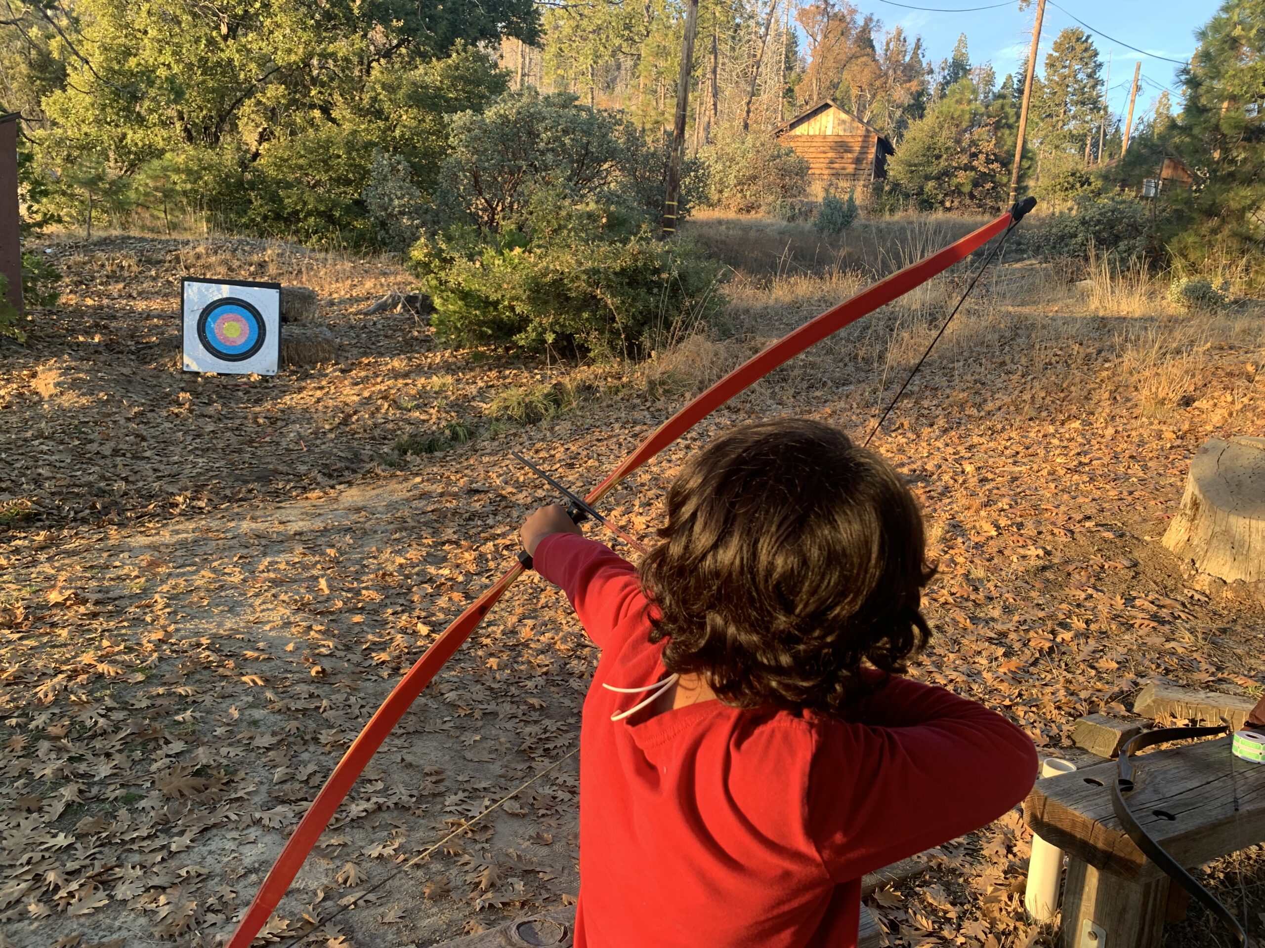 Young archer in red shirt drawing a bow, aiming at a target in a forest clearing with autumn leaves and a rustic cabin in the background.